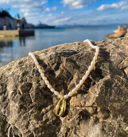 pearl necklace with gold cowrie shell pendant. Necklace is on a rock with blue sea and sky behind. 