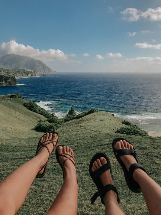 View looking over the sea from a clifftop. A couple dangle their feet in the view wearing flipflops