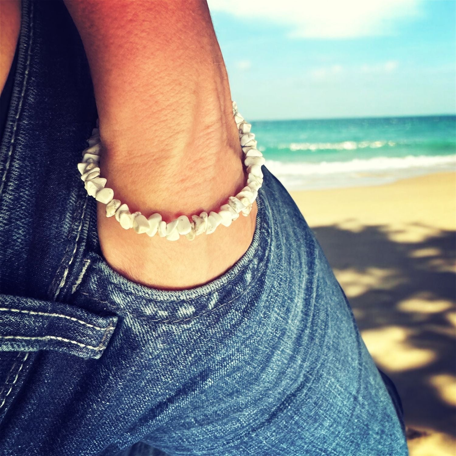 Person wearing jeans and a bracelet made of stone chips. Beach and sea is behind. 