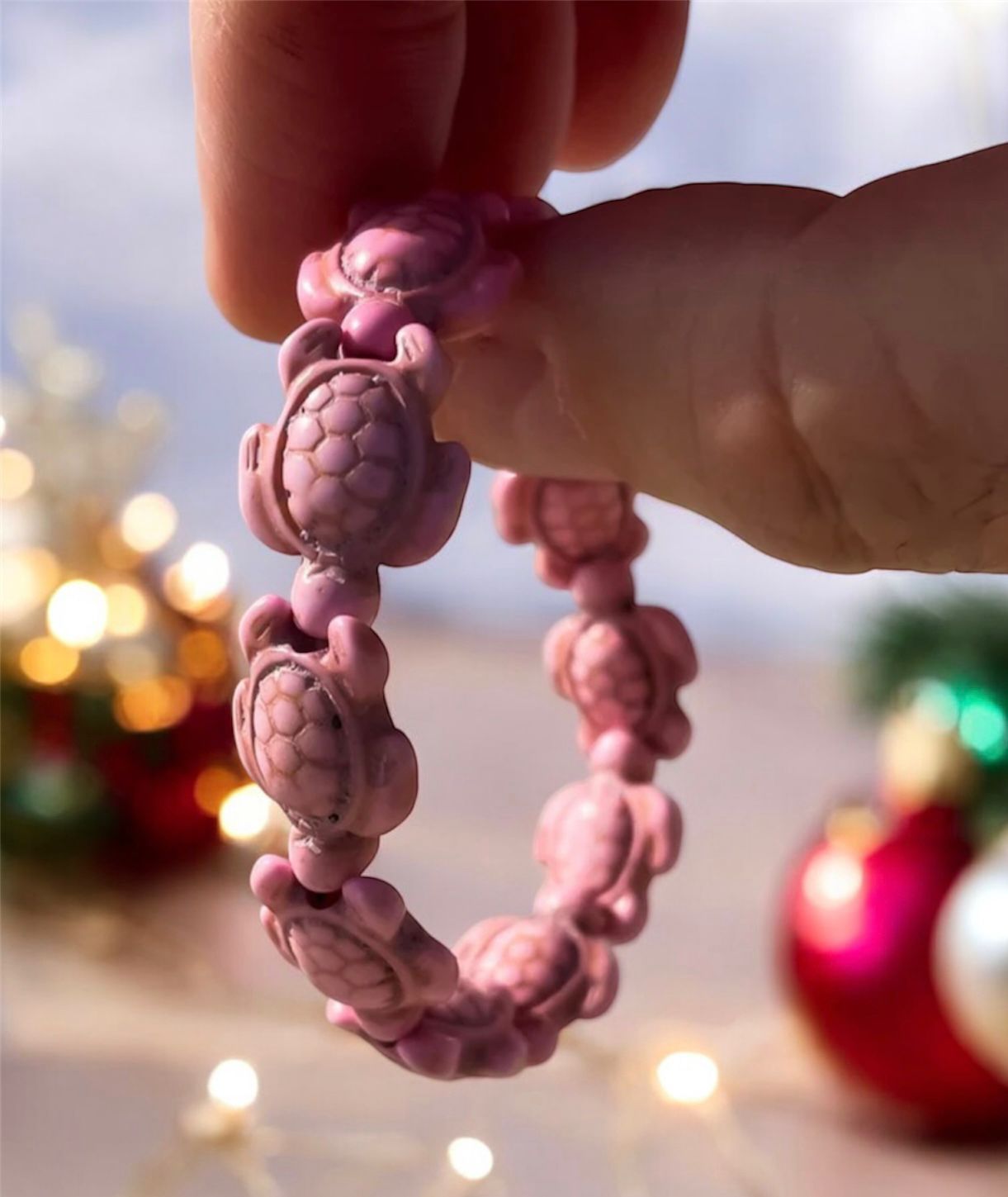 Someone holding up a pink sea turtle bracelet with Christmas decorations behind. 