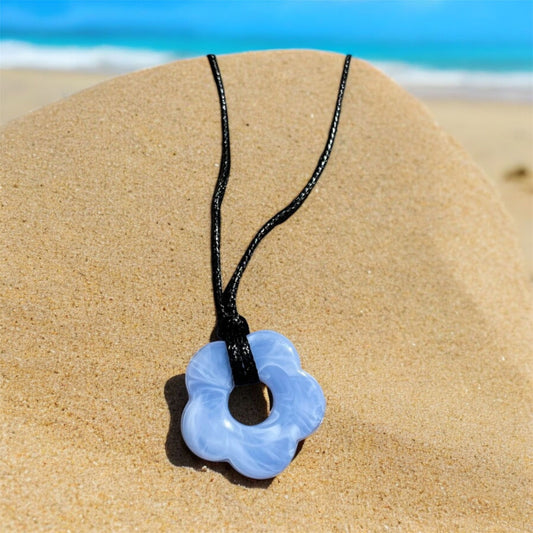 Black leather necklaces with blue flower shaped pendant. Necklace is on a beach with the sea behind. 