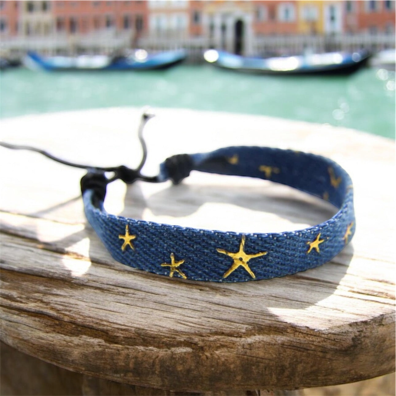 Blue denim friendship bracelet with gold stars on a wooden post with Venice canal scene behind. 