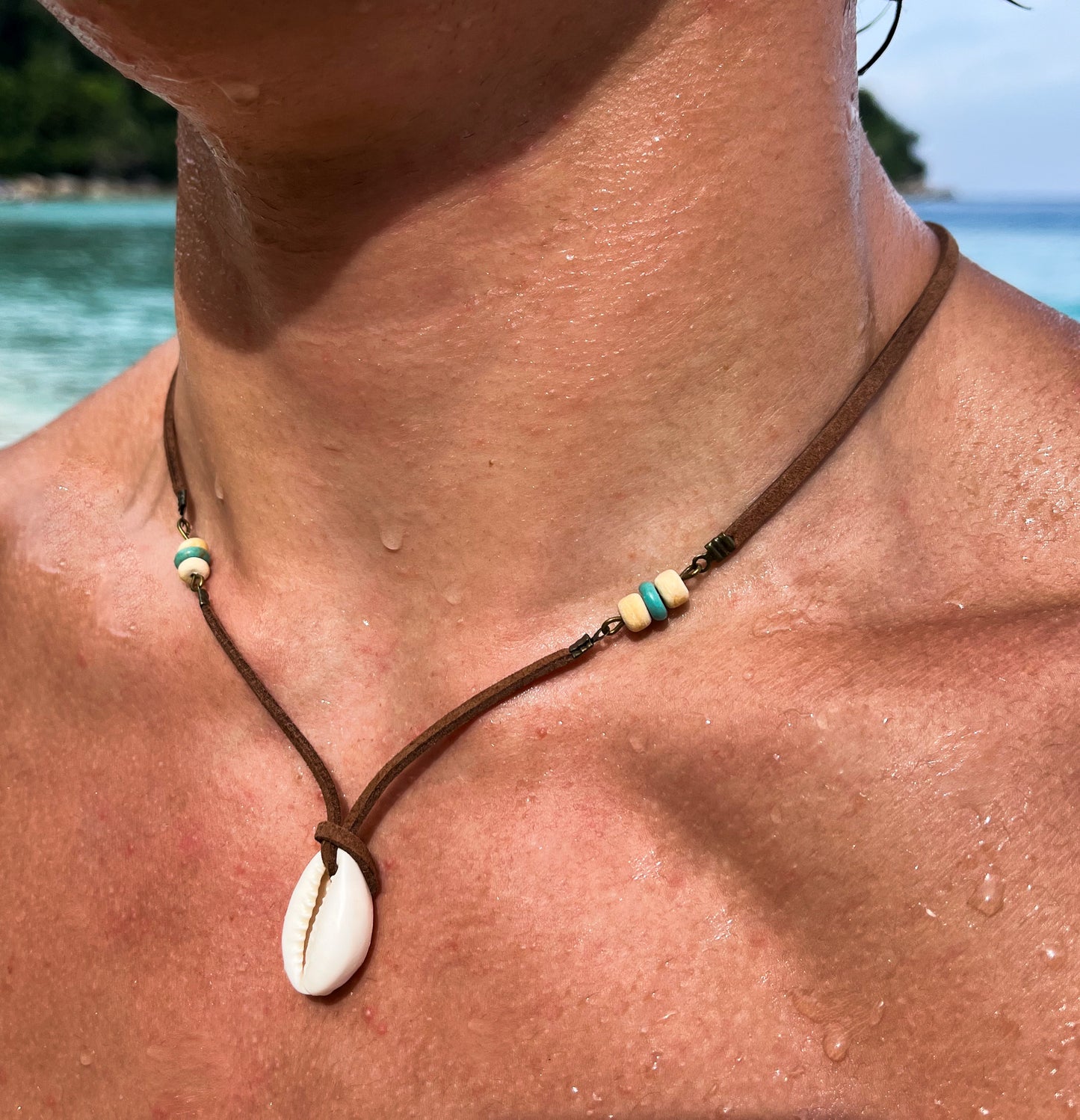 Model wearing a brown leather choker with cowrie shell pendant. Blue sea is behind. 