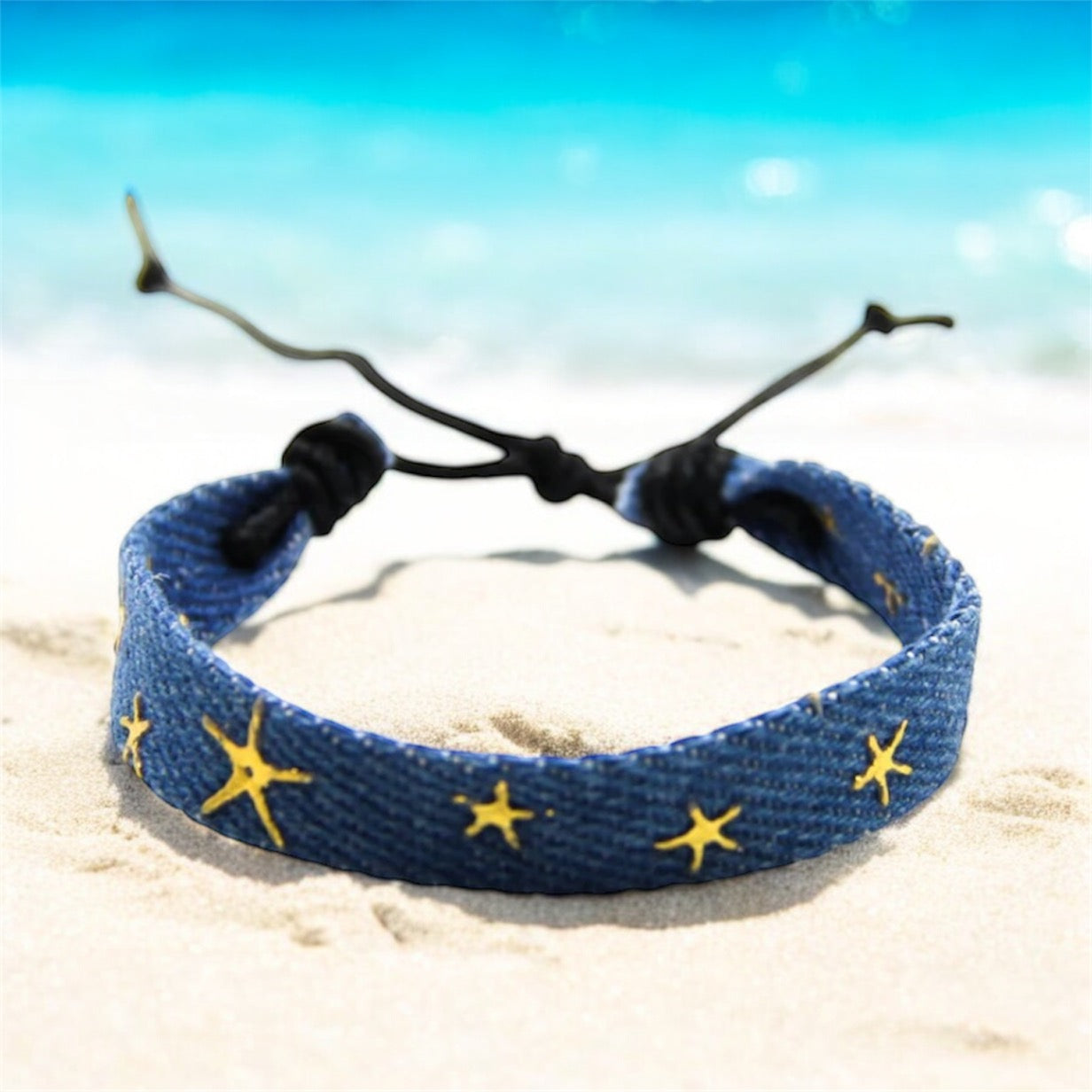 Blue denim bracelet with gold stars on a beach with blue sea behind. 