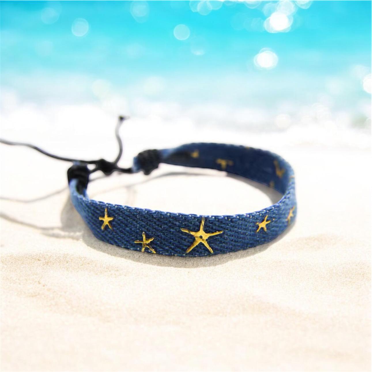 Blue denim friendship bracelet with gold stars on a beach with deep blue sea behind. 