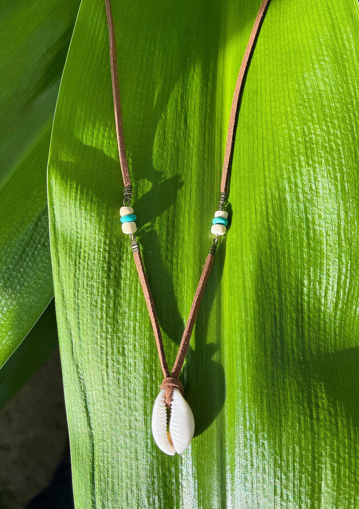 Brown leather necklace with shell pendant displayed on a tropical leaf