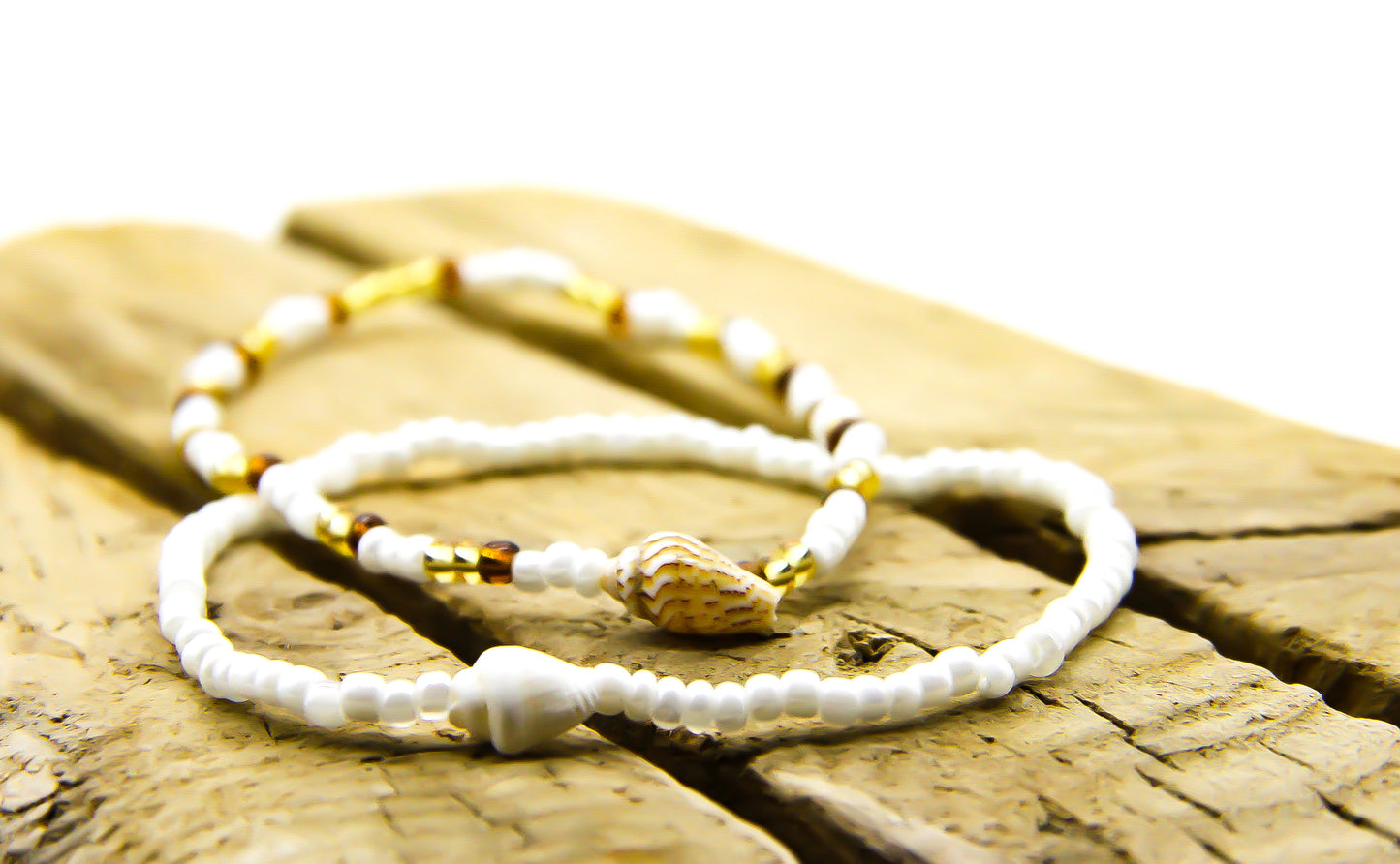 Close-up of 2 white and amber beaded bracelets with shell pendants on a piece of slatted wood