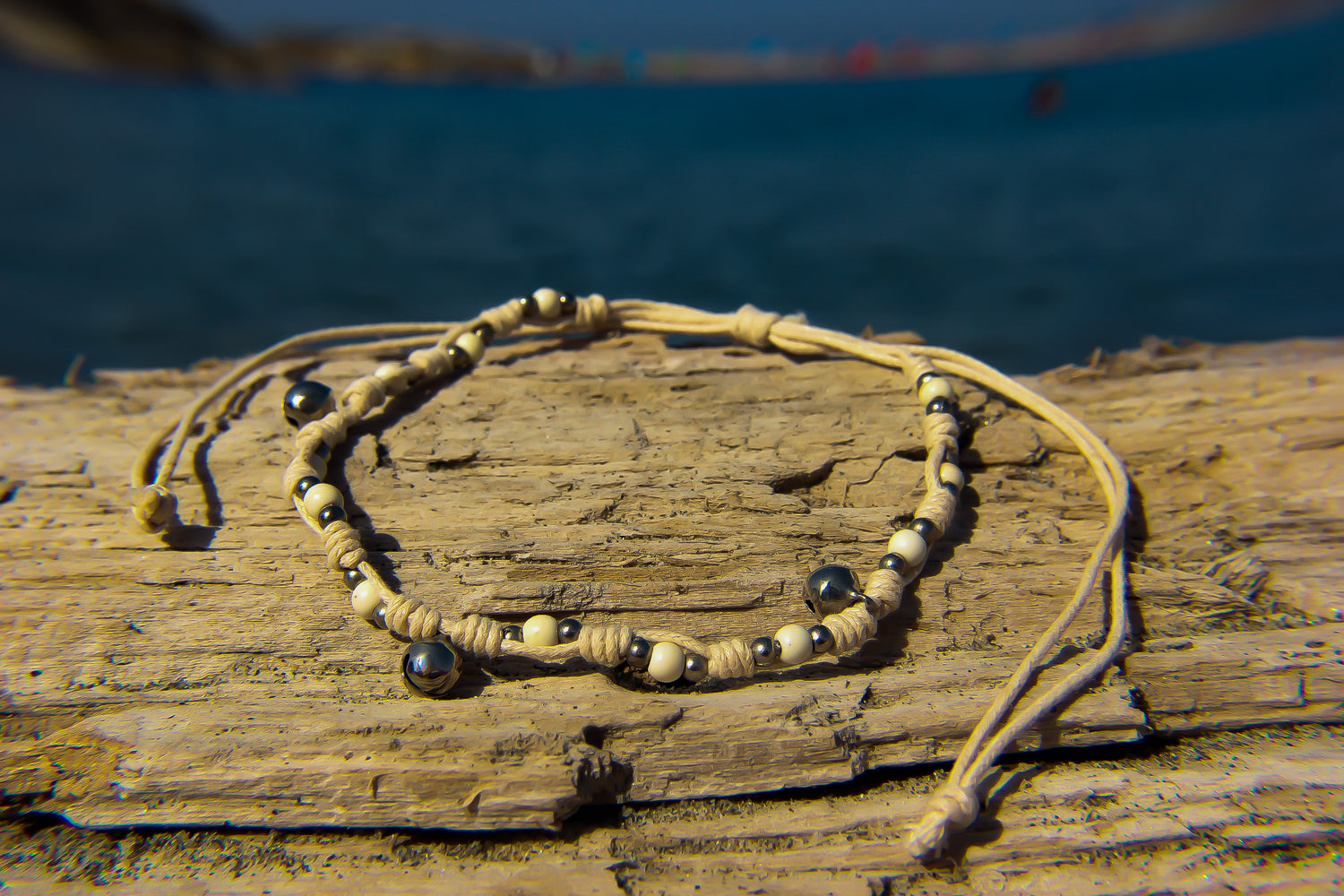 Close-up of string anklet with silver beads and bells | Anklet is on drift-wood in front of sea