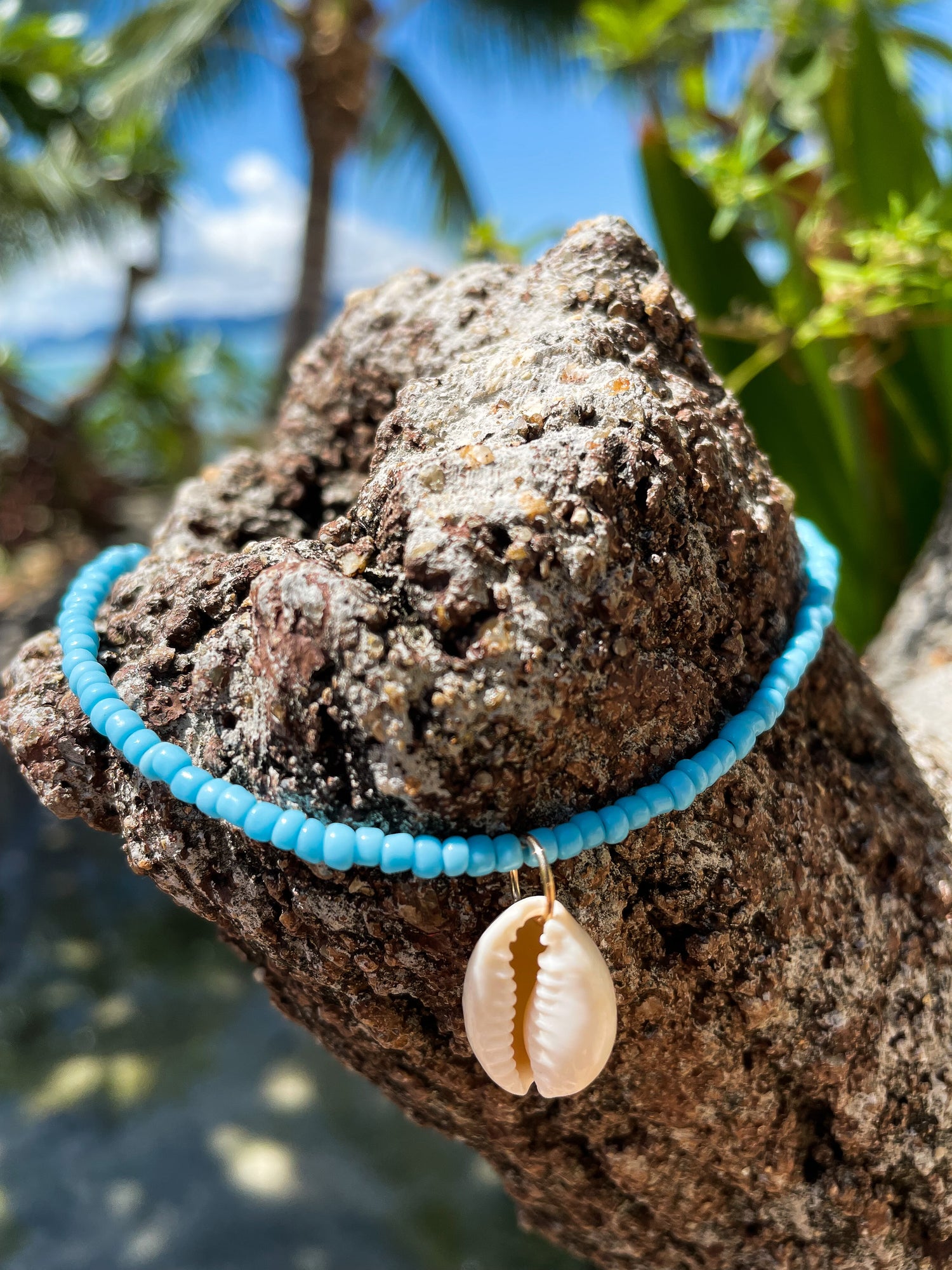 Blue seed beach choker with cowrie shell displayed in a beach setting with plams trees and sea behind. 