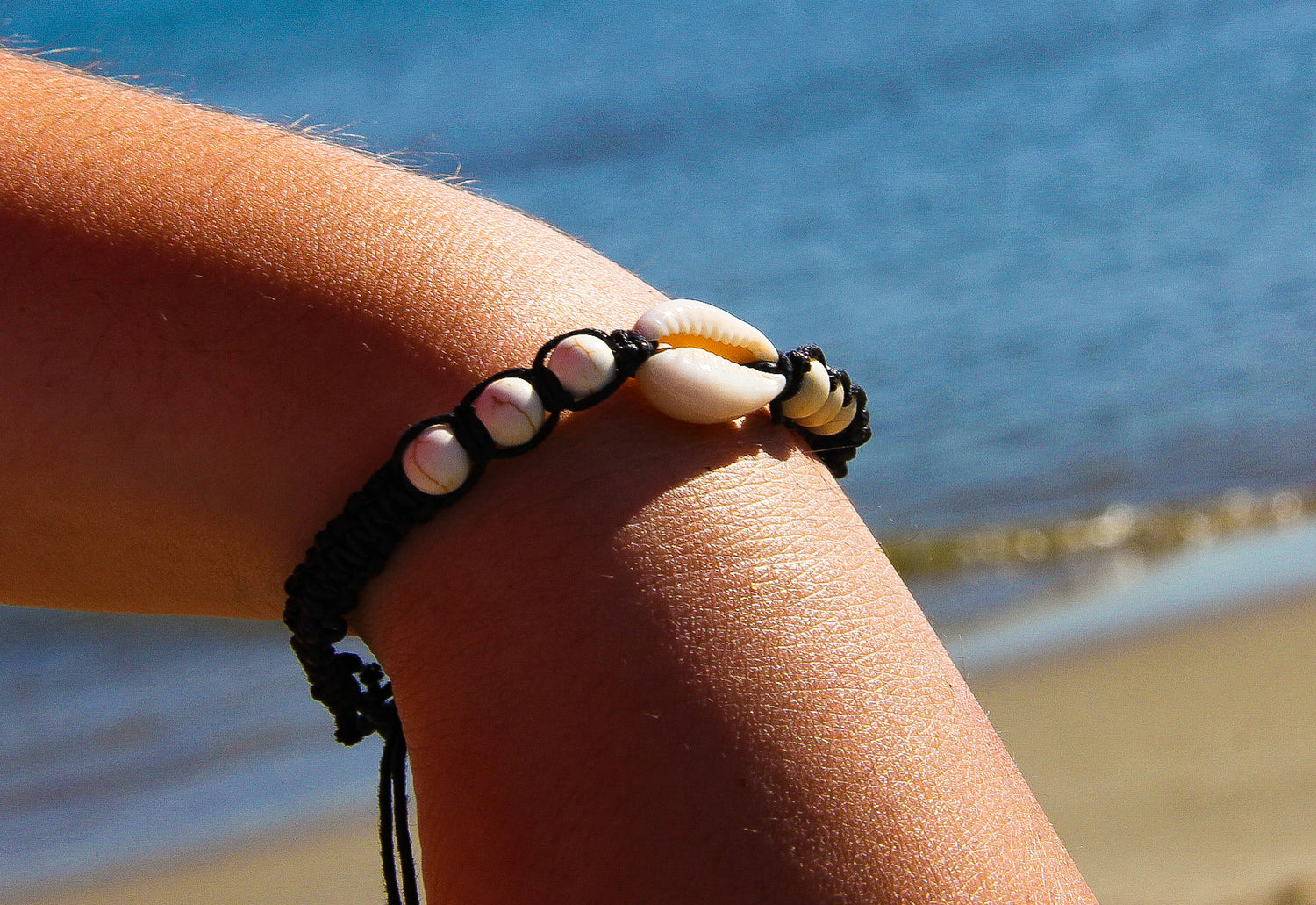 Braided black bracelet on model's wrist with white beads and cowrie shell, sea in background
