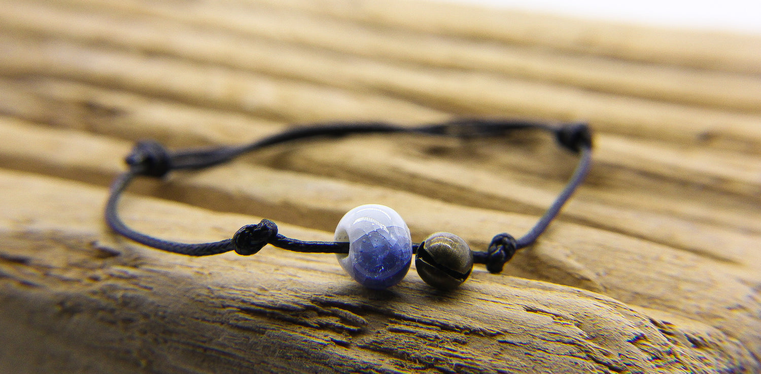 Black friendship bracelet with blue and white glass bead and brass bell. Bracelet is on a piece of wood. 