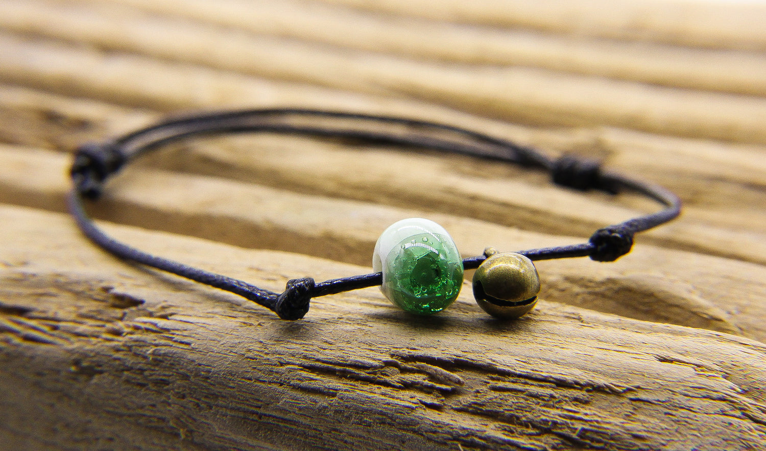 Black cord bracelet with green and white glass bead and tiny brass bell. Bracelet is displayed on a piece of slatted wood. 