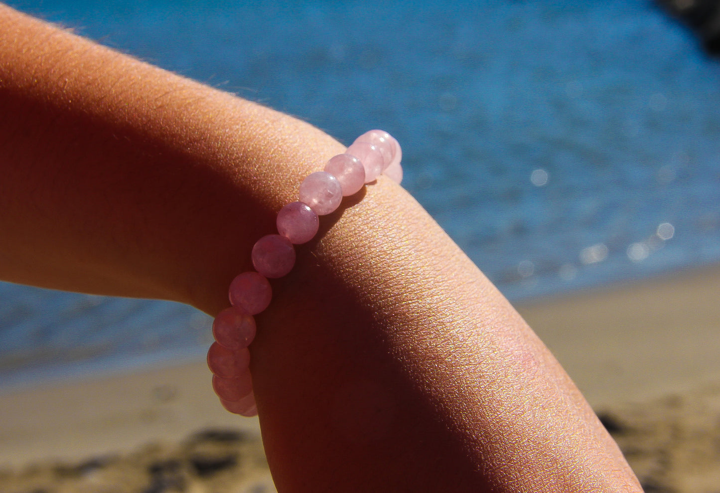 Close up of model's wrist wearing a pink gemstone beaded bracelet. Beach and blue sea in the background.