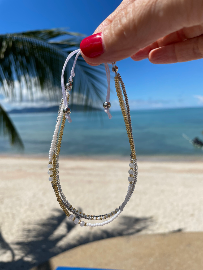 close up of model's hand holding white and gold beaded anklet with sea behind