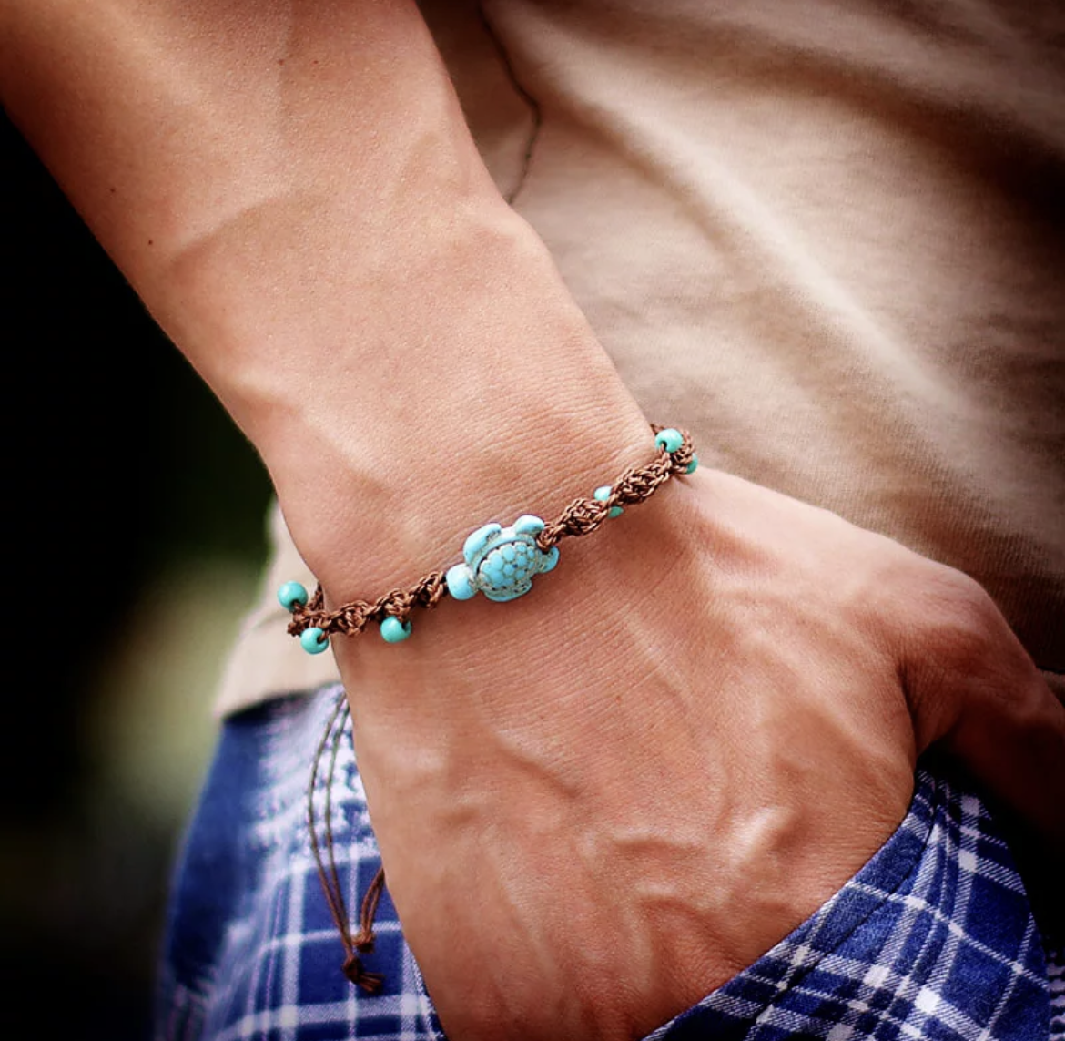 Close-up of male model's hand wearing brown woven friendship bracelet with turquoise sea turtle pendant