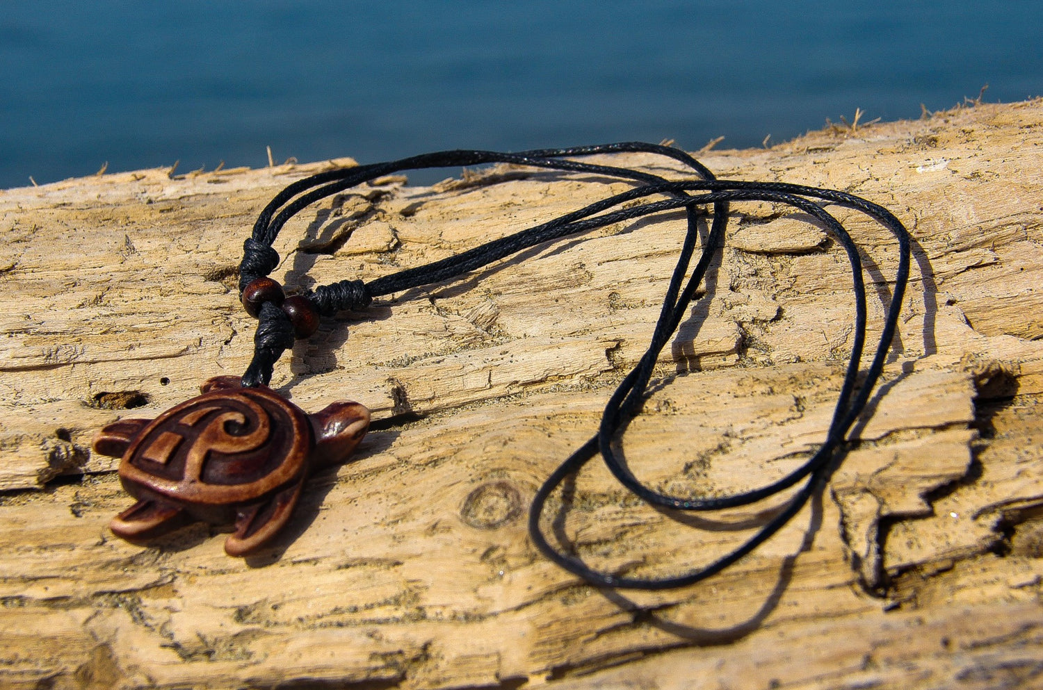 black cord necklace with carved wooden sea turtle pendant on a piece of wood with sea in background