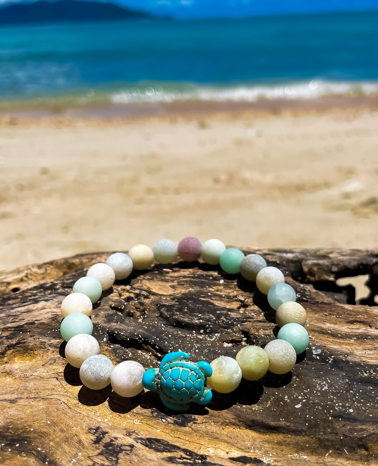 Stone bracelet with sea turtle pendant on driftwood with the ocean behind. Ben's Beach Jewellery 
